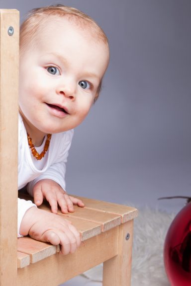 Ein lächelndes Kleinkind sitzt auf einem kleinen Stuhl neben einem roten Kugelornament.