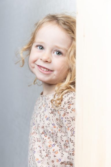 Lächelndes kleines Mädchen mit lockigem, blondem Haar, trägt ein blumiges Kleid.