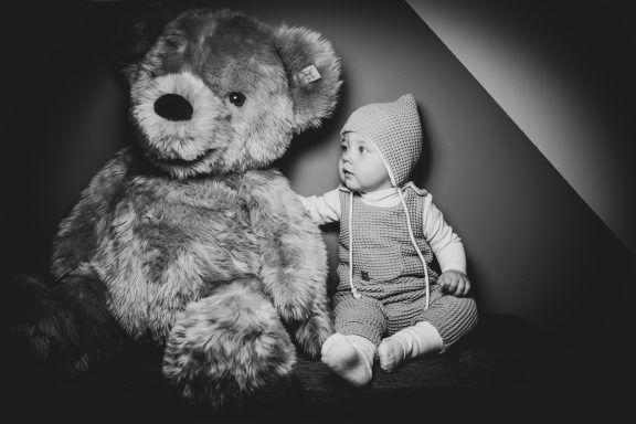 Ein Baby in einem gestrickten Hut sitzt neben einem großen Stoffbären.