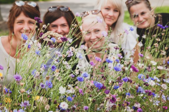 Fünf Frauen lächeln vor bunten Wildblumen.
