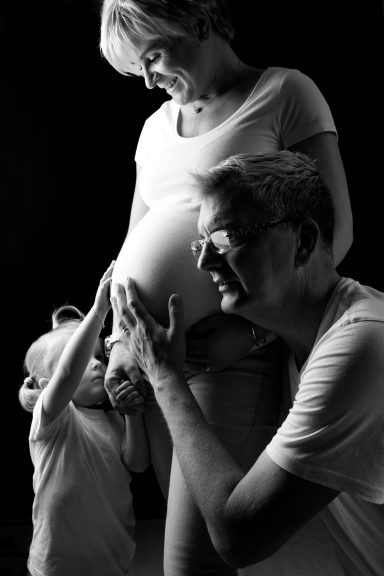 Eine schwangere Frau steht neben einem Mann und einem kleinen Kind, das den Bauch berührt.