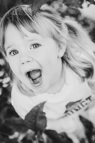 Lachendes Kind mit blonden Haaren, umgeben von grünen Blättern.