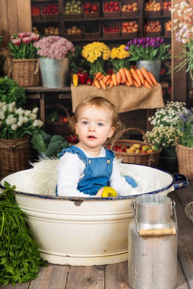 Ein Kleinkind sitzt in einer Schüssel neben frischem Gemüse und Blumen.
