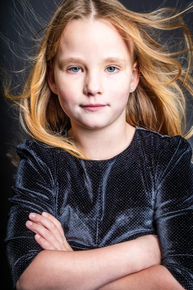 Ein junges Mädchen mit langen, fließenden Haaren und verschränkten Armen, ernst blickend.