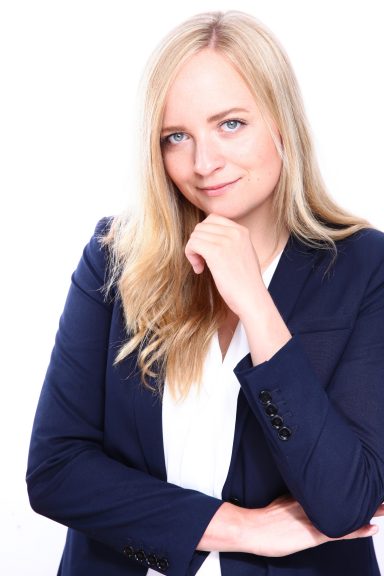 Junge Frau mit blonden Haaren in einem blauen Blazer, nachdenklich, leicht lächelnd.