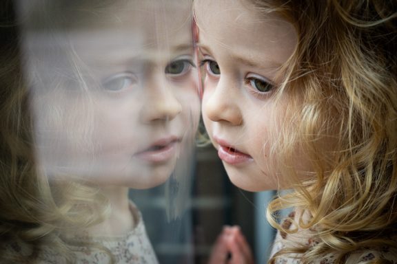 Ein Mädchen mit lockigem Haar schaut nachdenklich aus einem Fenster.