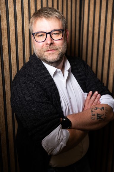 Mann mit Brille und Bart, gekleidet in einem weißen Hemd und schwarzem Überwurf, arm verschränkt.