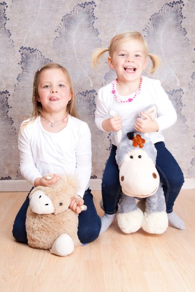 Zwei Mädchen sitzen auf Plüschtieren, lächeln und tragen weiße Oberteile.