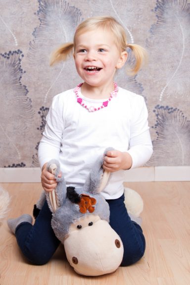 Ein Mädchen mit Zöpfen sitzt lächelnd auf einem Stoffpferd und trägt eine farbige Kette.