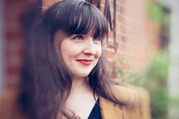 Lächelnde Frau mit langen, dunklen Haaren vor einer Backsteinmauer.