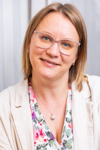 Lächelnde Frau mit Brille, blumigem Oberteil und heller Jacke, vor neutralem Hintergrund.