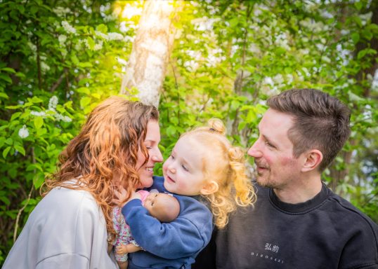 Familie mit Eltern und Kleinkind, umgeben von grüner Natur, lächelnd und glücklich.