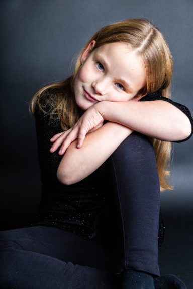 Ein Mädchen mit langen, blonden Haaren sitzt, lächelt sanft und trägt dunkle Kleidung.