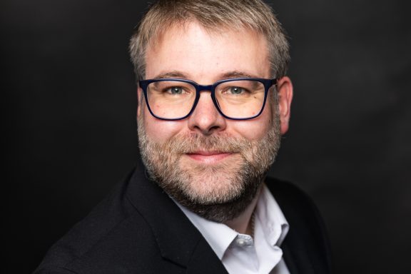 Mann mit Brille, Bart und Anzug, lächelt vor dunklem Hintergrund.