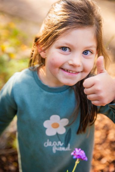 Ein Mädchen mit einem Daumen nach oben, trägt ein grünes Sweatshirt mit Blümchen.