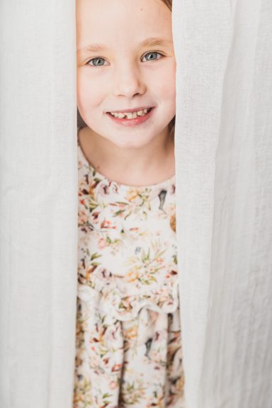 Lächelndes Kind mit floralem Kleid, das hinter einem weißen Vorhang hervorschaut.