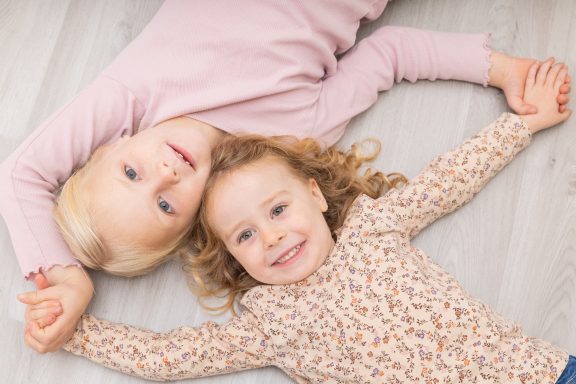 Zwei lächelnde Mädchen liegen auf dem Boden und halten sich an den Händen.