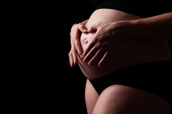 Hände halten einen runden, schwangeren Bauch vor dunklem Hintergrund.