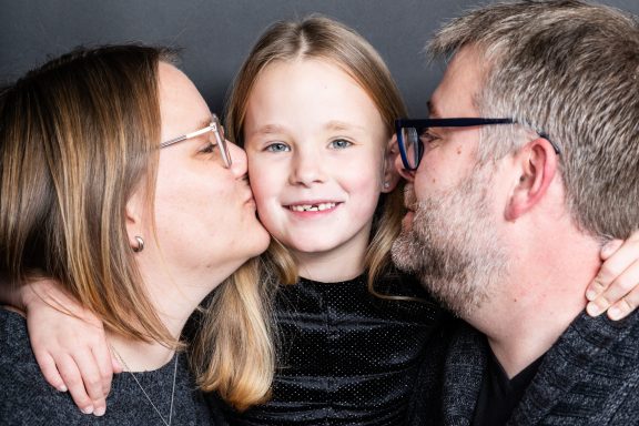 Familie posiert zusammen, ein Elternteil küsst das Kind, alle lächeln.