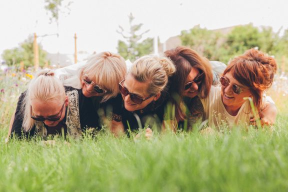 Fünf Frauen liegen im Gras und lachen, umgeben von Natur.