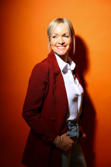 Frau mit blonden Haaren, lächelt in einem roten Blazer vor orangefarbenem Hintergrund.