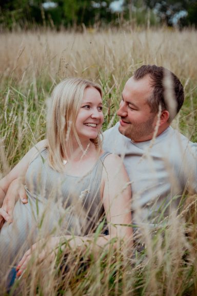 Paar sitzt in hohem Gras und lächelt sich an, eine entspannte, liebevolle Stimmung.