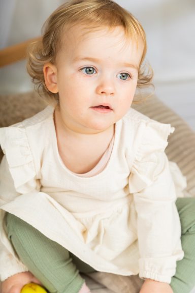 Kleines Mädchen mit blonden Locken, hellem Kleid und grünen Leggings, schaut nachdenklich.