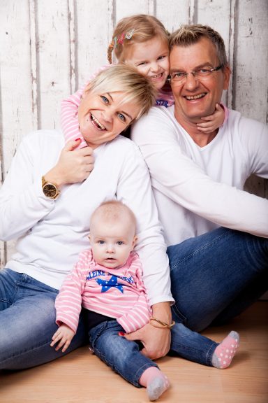 Familie mit zwei kleinen Kindern, sitzen lächelnd auf dem Boden vor einer Holzpaneelwand.