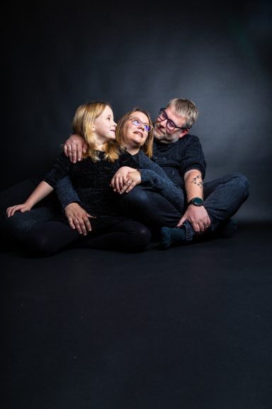 Drei Personen in einer entspannten Positur, umarmend auf dem Boden sitzend.