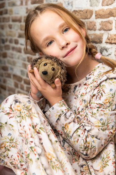 Ein Mädchen mit Zopf hält ein Stofftier in Form eines Igels umarmt, vor einer Ziegelwand.