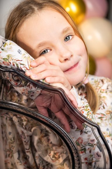 Ein lächelndes Mädchen mit blonden Haaren sitzt auf einem Stuhl, umgeben von farbigen Ballons.