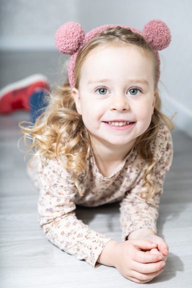 Ein lächelndes Kind mit Locken und einem rosa Haarband liegt auf dem Boden.