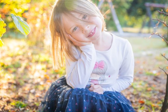 Ein lächelndes Mädchen sitzt im Freien zwischen bunten Blättern und strahlt Freude aus.