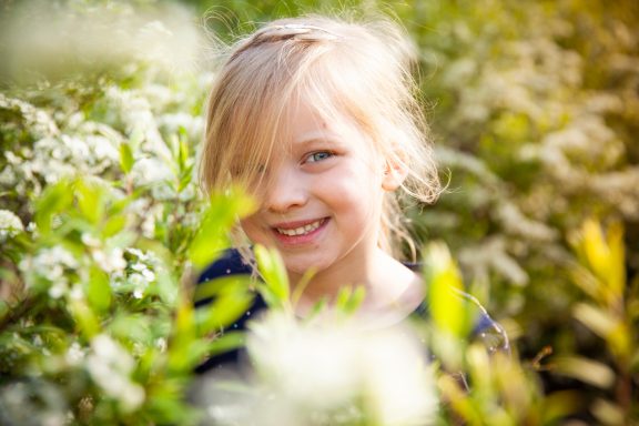 Ein lächelndes Mädchen versteckt sich zwischen grünen Pflanzen und Blumen.