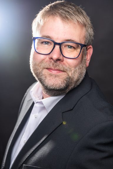 Mann mit Brille und Bart in Anzug, lächelnd vor dunklem Hintergrund.