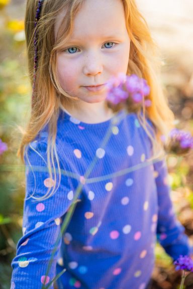 Ein kleines Mädchen mit langen, blonden Haaren und blauer gepunkteter Bluse, umgeben von Blumen.