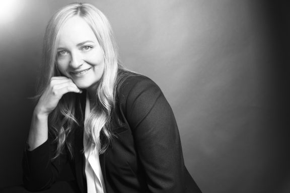 Frau mit langen Haaren in einem Anzug, lächelt und stützt den Kopf auf ihre Hand.