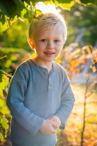 Ein lächelnder Junge mit blonden Haaren steht zwischen Pflanzen im Sonnenlicht.