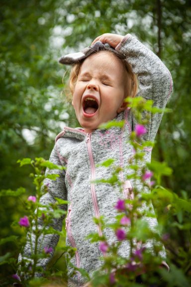 Lachendes Mädchen mit grauem Pullover steht zwischen grünen Pflanzen und lila Blumen.
