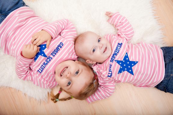 Zwei Kinder in gestreiften T-Shirts mit blauen Sternen liegen auf einem hellen Teppich.