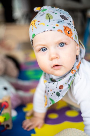 Baby mit blauen Augen, trägt ein buntes Tuch, liegt auf einer Spielmatte.