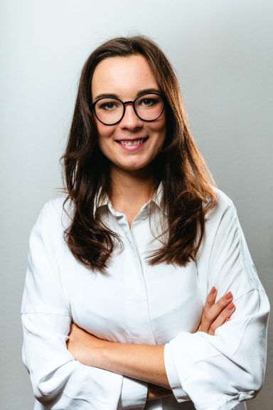 Junge Frau mit Brille, lächelt und hat die Arme vor der Brust verschränkt.