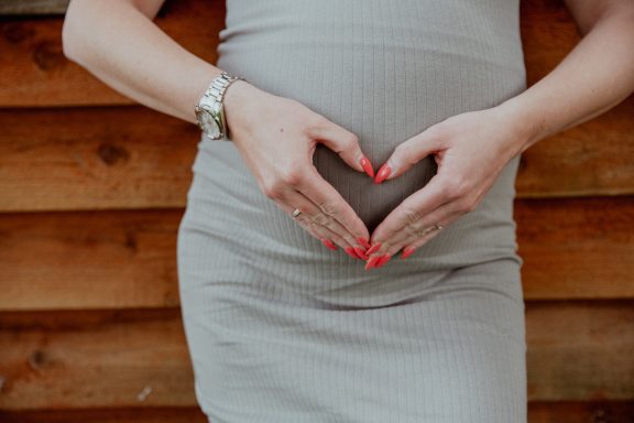 Schwangere Person formt mit Händen ein Herz auf dem Bauch.