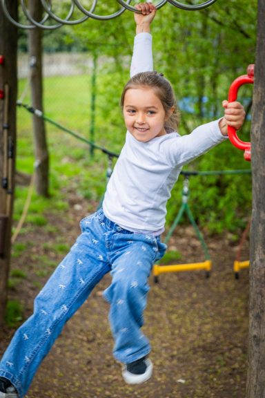 Mädchen schwingt an einer Kletterstange im Spielplatz, lächelt dabei.