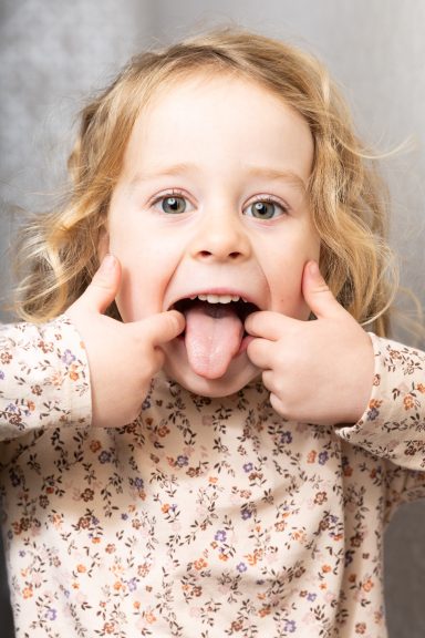 Ein Kind mit lockigem Haar zieht die Zunge heraus und zeigt die Daumen nach oben.